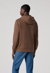 Felpa marrone con tasca a marsupio frontale e cappuccio regolabile, realizzata in tessuto morbido, abbinata a jeans in denim blu.