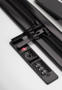 Cremallera negra con dos lengüetas de extracción, que presenta un candado de combinación. Base de plástico duro con un logotipo de TSA en rojo y discos numéricos claros.