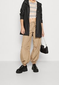 Czarna wodoodporna kurtka z kapturem, paskowany krótki top, beżowe luźne spodnie oraz czarne masywne buty na platformie, trzyma małą czarną torbę.