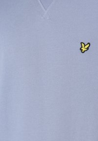 Ljust blå V-ringad tröja gjord av mjukt, strukturerat tyg. Har en liten svart och gul logotyp av en fågel på vänster bröstområde.