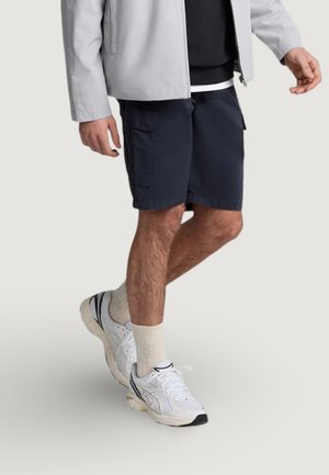 Persona que lleva pantalones cortos cargo negros, zapatillas blancas, calcetines beige, una camiseta negra y una chaqueta gris claro caminando sobre un fondo liso.