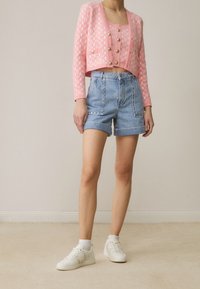 Różowy krótki kardigan z geometrycznym wzorem i złotymi guzikami, zestawiony z jasnoniebieskimi szortami dżinsowymi ozdobionymi srebrnymi ćwiekami i podwiniętymi nogawkami. Białe sneakersy dopełniają całość.