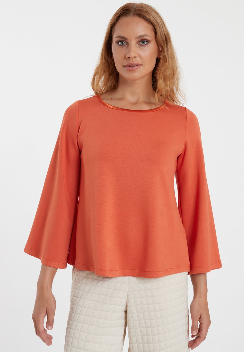 Orange Langarmshirt mit runder Halslinie und Glockenärmeln; weicher Stoff. Kombiniert mit cremefarbenen, gesteppten Hosen.