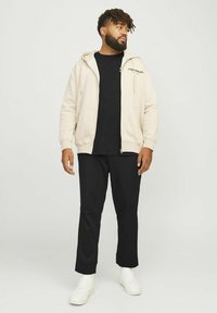 Haut de survêtement zippé beige clair, t-shirt noir, pantalon noir et baskets blanches. Tissu lisse et coupe décontractée avec logo brodé sur le hoodie.