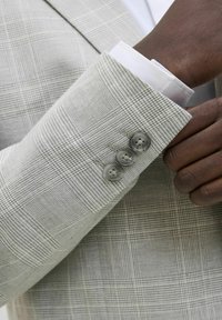 Blazer gris claro con un patrón de cuadros texturizado, tres botones grises en la manga y un cuello de camisa blanca asomando por debajo de la manga.