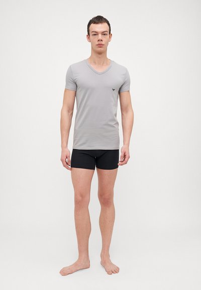 Sivi v-neck majica od rastezljivog materijala, u paru s crnim kratkim hlačama. Ima kratke rukave i mali logo na grudima.