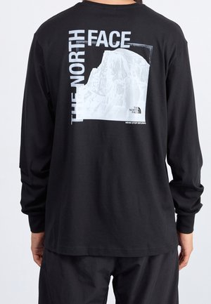 Schwarzes Langarmshirt mit weißem Bergmotiv und dem Schriftzug "THE NORTH FACE" auf dem Rücken, getragen von einer Person, die von der Kamera weg schaut.