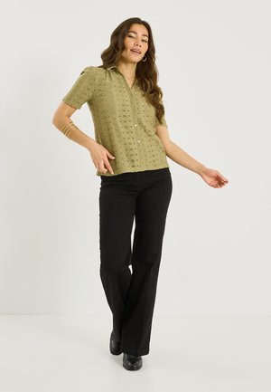 Femme portant un chemisier à manches courtes boutonné vert olive avec motif en dentelle, pantalon large noir, chaussures noires et bracelets dorés au bras gauche.