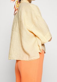 Person iført en løs, lys gul linnedskjorte med ærmer til albuen og skarpe orange, tætsiddende bukser, med et flerfarvet tørklæde knyttet i håret.