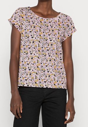Vrouw draagt een blouse met korte mouwen met een bloemmotief in roze, geel en zwart, gecombineerd met een zwarte broek.