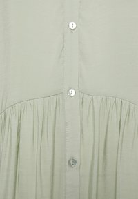 Helles grünes Blusenshirt mit Knopfleiste, gefaltetem Detail in der Taille und glänzenden Knöpfen. Glatter, fließender Stoff mit weicher Textur.