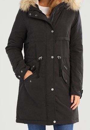 Žena v černém zimním kabátě délky po kolena s ozdobou z umělé kožešiny na kapuci, předními knoflíky, zipem, stahovacími šňůrkami a několika předními kapsami.