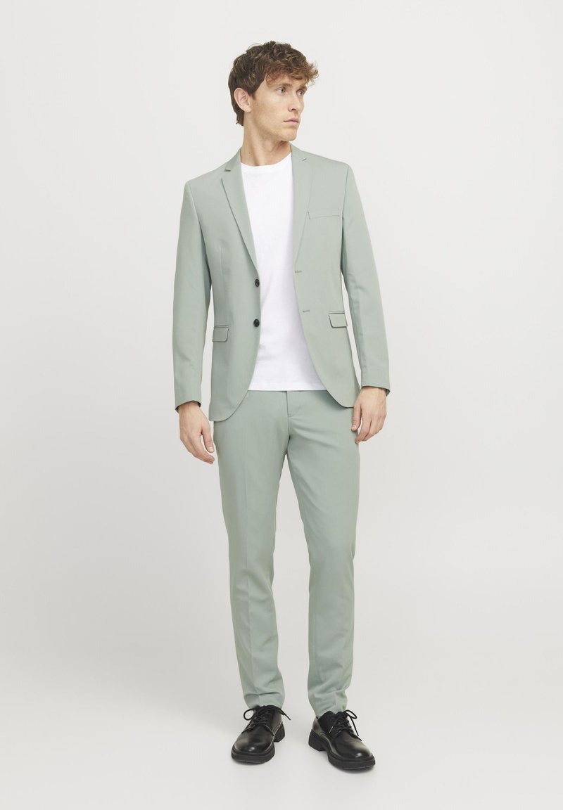 Lys grønn dress med slim fit-design; blazer har spisse revers og lommer; kombinert med en hvit t-skjorte og svarte sko.