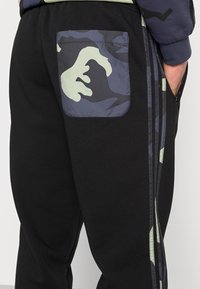 Pantalones de chándal negros con un bolsillo de camuflaje, que presentan una tela texturada y rayas en contraste a los lados en verde y azul marino.