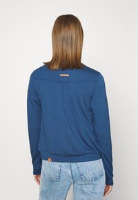 Blå långärmad topp i mjukt material, med rundad halslinning, veck bak och en liten märkesetikett vid halsen och fållen.