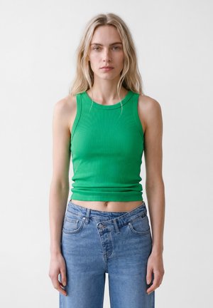 Blonde Frau trägt ein grünes ärmelloses, geripptes Crop-Top und hoch taillierte blaue Jeans, wobei der oberste Knopf ungleichmäßig geschlossen ist.