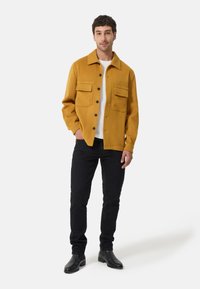 Giacca color senape con texture liscia, dotata di due tasche sul petto e bottoni neri, abbinata a pantaloni neri slim-fit e stivali neri.