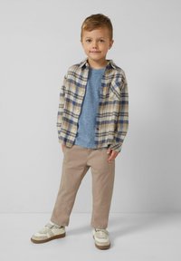 Junger Junge in beiger kariertem Hemd über einem blauen T-Shirt, khakifarbener Hose und weißen Sneakern, der vor weißem Hintergrund steht.