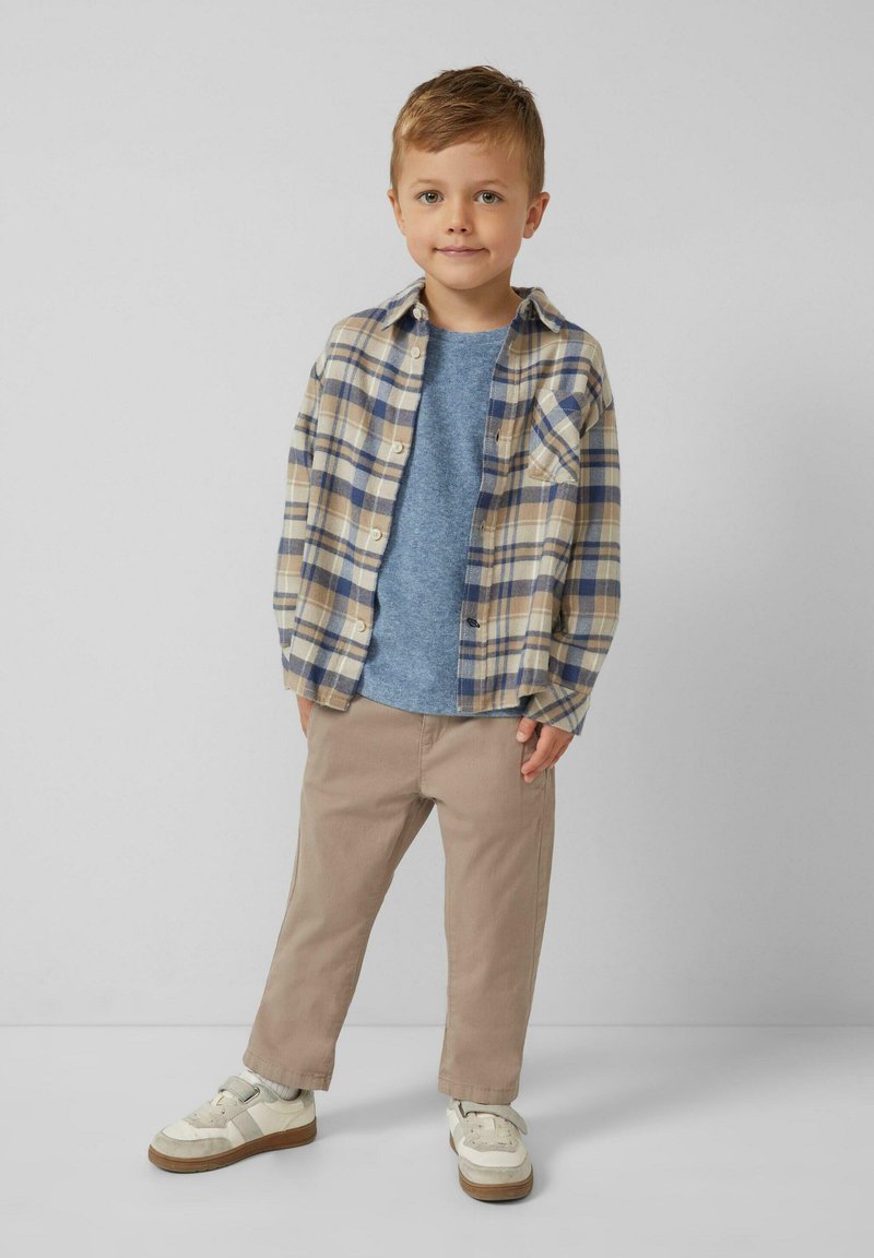 Junger Junge in beiger kariertem Hemd über einem blauen T-Shirt, khakifarbener Hose und weißen Sneakern, der vor weißem Hintergrund steht.