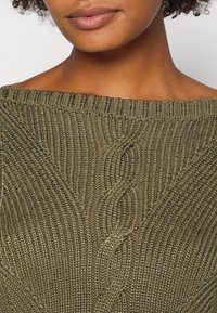 Gros plan d'une personne portant un pull tricoté vert olive avec un motif tressé au centre et un large col au-dessus de la clavicule.