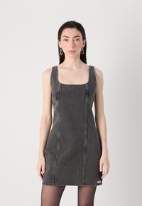 En ung kvinne med langt hår har på seg en ermeløs, mørk denimkjole. Hun står foran en lys grå bakgrunn og viser et selvsikkert uttrykk.