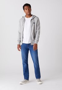 Szara bluza z kapturem i zamkiem, noszona na białym T-shircie, w połączeniu z niebieskimi dżinsami i białymi trampkami. Gładki materiał, minimalistyczny design.