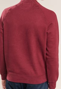 Person wearing a plain long-sleeve red sweatshirt viewed from the back, paired with dark blue pants.