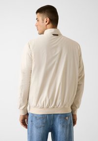 Hellbeige Bomberjacke mit glatter, glänzender Textur, gerippten Bündchen und hohem Kragen, mit einem kleinen Logo-Patch am Hals.