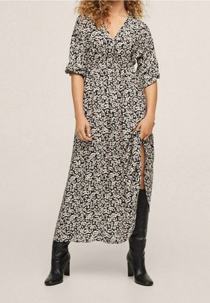 Kobieta w czarno-białej sukience midi w kwiaty z podwiniętymi rękawami i czarnych, wysokich kozaczkach na obcasie, trzymająca rozcięcie sukienki otwarte.