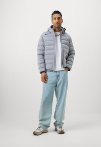 Grå oppustelig jakke med vandret quiltet mønster, lynlås foran og hætte. Båret over en hvid t-shirt, sammen med lyseblå jeans og sneakers.