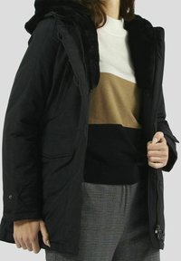 Kvinna i en svart huvtröja med pälsdetaljer, färgblockerad tröja i vitt, beige, svart och grå rutiga byxor. Hand håller kappan öppen.