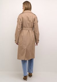 Trench coat beige con vita cintura, spacco posteriore e dettagli con bottoni. Indossato sopra jeans azzurri chiari e scarpe con tacco marroni.
