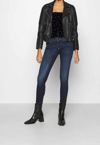 Svart läderbikerjacka med ribbade axlar, bärs över en svart samettopp, kombinerad med mörkblå skinny jeans och svarta klackstövlar.