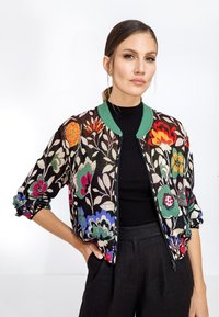 Giacca bomber con ricamo floreale multicolore, colletto e polsini a coste verdi, chiusura con zip e fondo nero. Indossata sopra una maglietta nera.