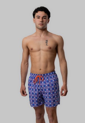 Giovane uomo in piedi senza maglietta che indossa pantaloncini da bagno blu a fantasia con coulisse arancione, su uno sfondo grigio uniforme.