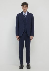 Mann steht frontal und trägt einen marineblauen, gestreiften Dreiteiler, ein gestreiftes Hemd, eine gestreifte Krawatte und schwarze Schuhe auf einem weißen Hintergrund.