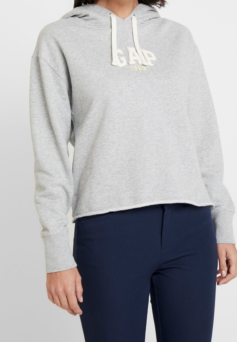Sudadera gris con capucha y un diseño recortado, que presenta un logo frontal de GAP en amarillo y detalles de cordones. Tejido suave y texturizado.