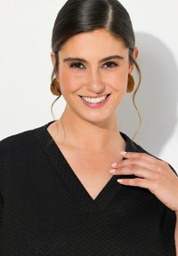 Top nero e texturizzato con design a V. Capelli lisci, orecchini leggeri e trucco naturale. Mano appoggiata delicatamente sul colletto.