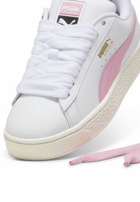 Baskets blanches Puma Suede avec une bande rose, lacets blancs noués et une paire supplémentaire de lacets roses à côté de la chaussure.