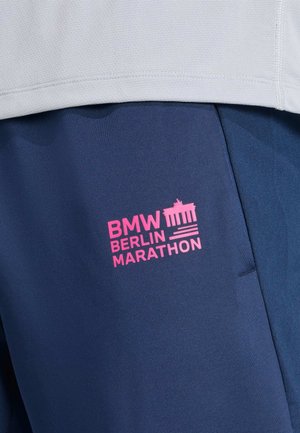 Nahaufnahme von marineblauen Sporthosen mit pinkem BMW Berlin Marathon-Logo und einem weißen strukturierten Shirt darüber.