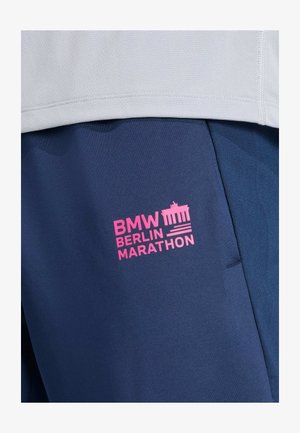 Nahaufnahme von marineblauen Sporthosen mit pinkem BMW Berlin Marathon-Logo und einem weißen strukturierten Shirt darüber.