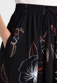 Černá sukně s elastickým pasem a šňůrkou, zdobená velkými bílými, červenými a hnědými květinovými a listovými vzory, ruka spočívající v kapse.