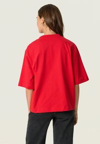 Red cotton t-shirt with short sleeves, a crew neck, and a loose fit, displayed from the back. Fabric has a smooth texture.