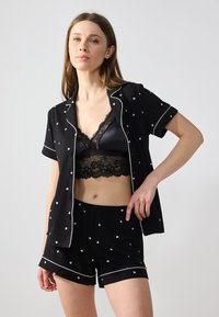 Svart kosesett med kortermet topp og shorts med hvite hjerte-mønstre. Toppen har kontrasterende hvite kanter, og en blondet bralette er til brukt under.