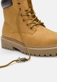 Beige Knöchelstiefel mit strukturiertem Obermaterial, robustem Sohlenprofil und gelb-braunen Schnürsenkeln mit einem markierten Metallanhänger. Glatte Ferse als Akzent.