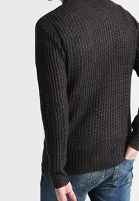 Donker gebreide trui met een gestructureerd patroon, voorzien van lange mouwen en een geribde zoom, gecombineerd met blauwe spijkerbroek.