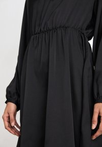 Black dress with long sleeves and gathered waist. Made from a smooth, matte fabric. Features a relaxed fit and slightly puffed sleeve cuffs.