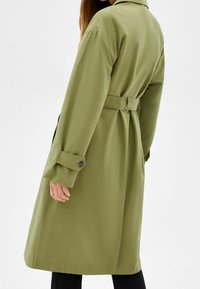 Olivgrön trenchcoat med bälte i midjan, långa ärmar och knäppningar vid ärmsluten. Mjuk tygkvalitet med en slät textur, fallande till knälängd.