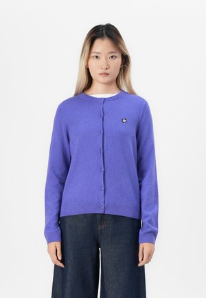 Donna con capelli lunghi e lisci, che indossa un cardigan viola con i bottoni e jeans scuri a gamba larga, in piedi davanti a uno sfondo bianco semplice.