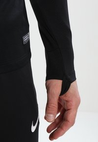 Schwarz langärmliges Sportoberteil mit taillierter Passform, ausgestattet mit einem Daumenloch am Bündchen und einem kleinen Markenetikett an der Seite.